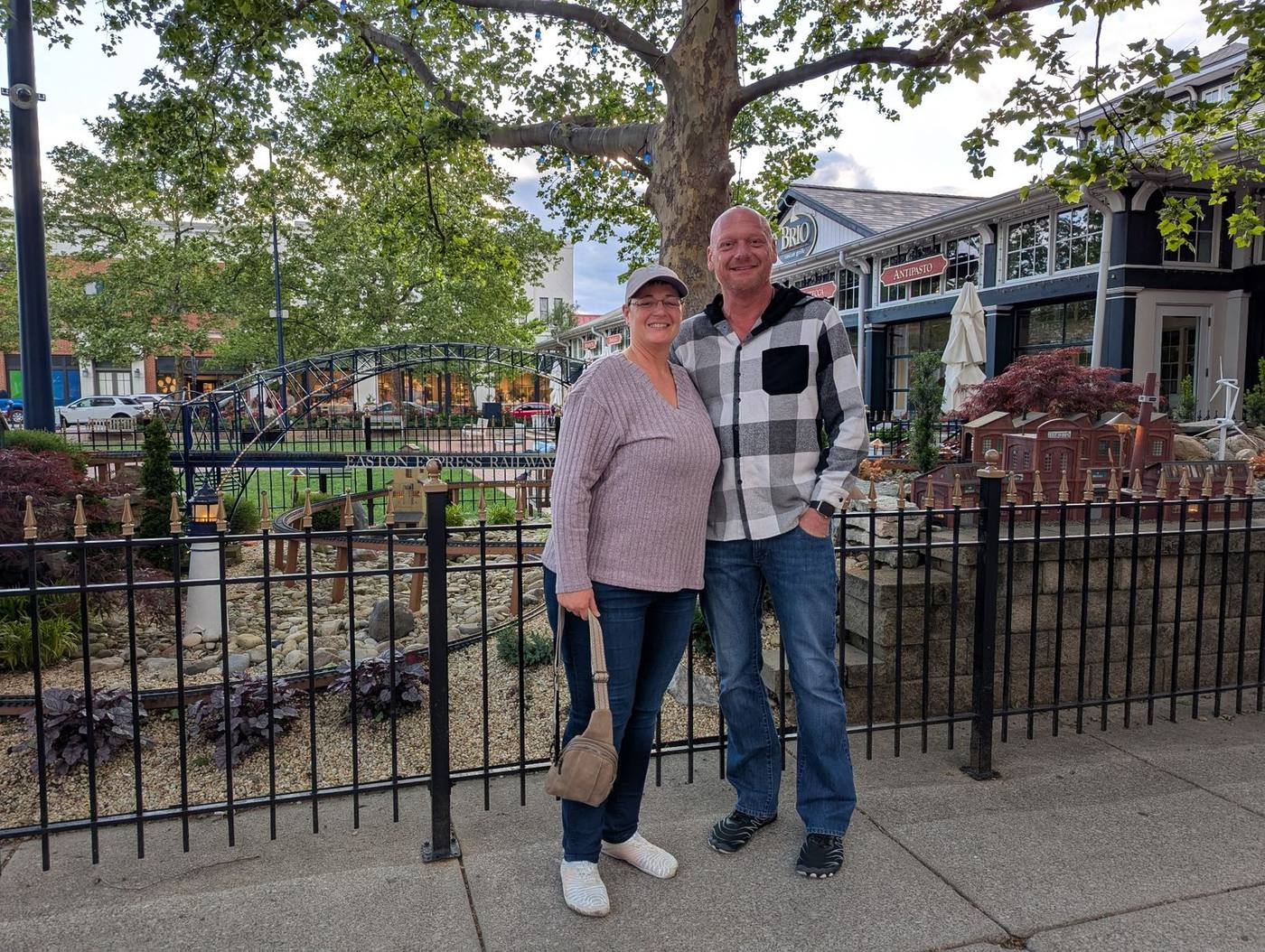 Stan and his wife standing together outdoors on a sidewalk near a small garden railroad.