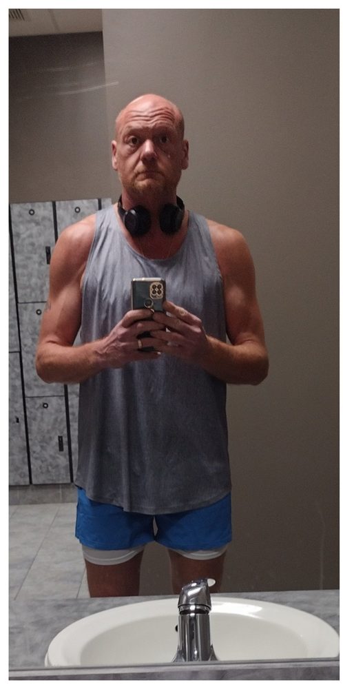 A later photograph of Stan in a gym mirror, wearing a tank top and holding a phone.